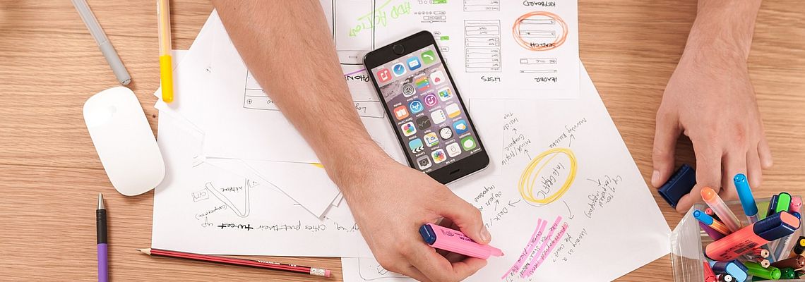 Hands, next to a smartphone, drawing differently coloured graphics on white paper sheets